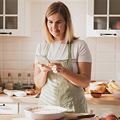 A woman in an apron uses a smartphone in a kitchen with wooden cabinets and a tiled backsplash.